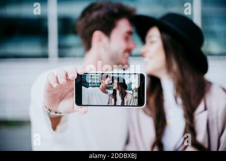 Smiling man taking photograph through smart phone while looking at female partner in city Stock Photo