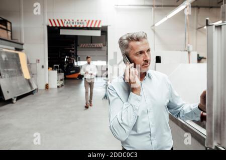 Businessman talking on mobile phone while colleague walking on background at industry Stock Photo