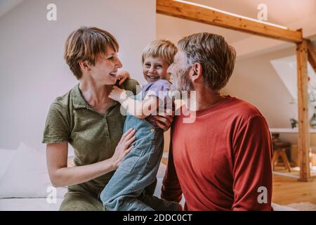 Happy mother cuddling son while father sitting beside on bed at home Stock Photo