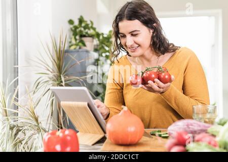 Smiling woman holding tomato while using digital tablet at home Stock Photo
