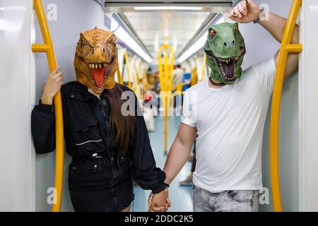 Male and female friends holding hands while wearing dinosaur mask in train Stock Photo