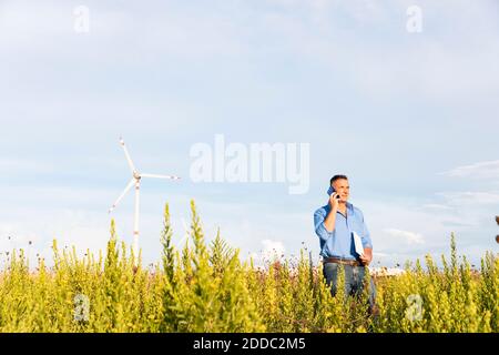Male engineer talking through mobile phone and document while standing against wind turbines on field Stock Photo