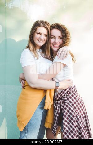 Smiling female friends hugging each other against wall Stock Photo