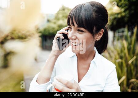 Smiling female engineer holding coffee cup while talking on mobile phone Stock Photo