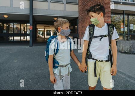 Brothers wearing protective face mask holding hands standing on street in front of school building Stock Photo