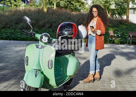 Smiling woman holding helmet on motor scooter container while using smart phone during sunny day Stock Photo