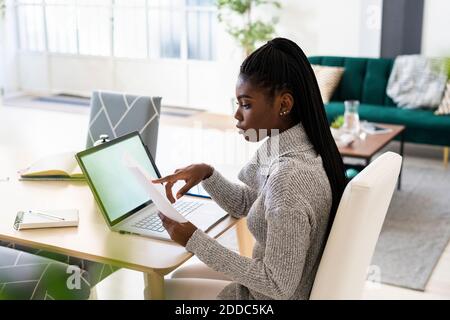Female student reading paper while studying on laptop at home Stock Photo