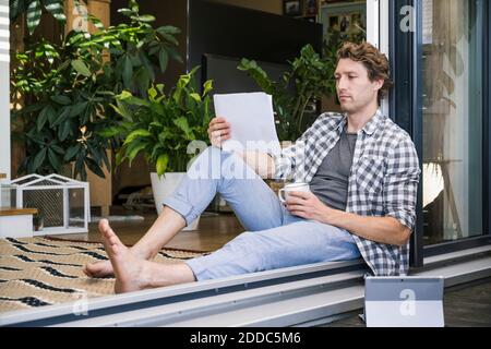 Mid adult man with coffee cup holding paper while sitting at home Stock Photo