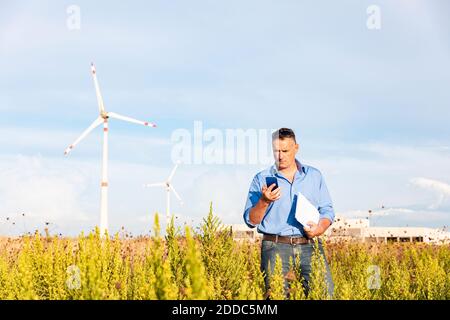 Mature businessman holding smart phone and document while standing against wind turbines on field Stock Photo