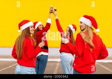 Happy young women in santa claus hats, friends cheerfully celebrate new year and drink drinks in glass bottles, on the background of a yellow wall out Stock Photo