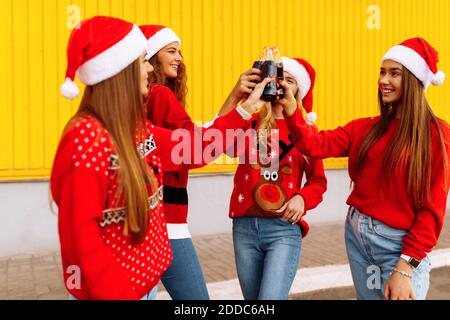 Happy young women in santa claus hats, friends cheerfully celebrate new year and drink drinks in glass bottles, on the background of a yellow wall out Stock Photo