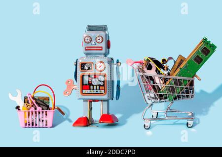 Studio shot of vintage robot toy standing between miniature shopping cart and basket filled with electronic equipment Stock Photo