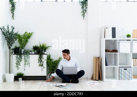 Young businessman analyzing documents while sitting with laptop on floor against white wall in office Stock Photo