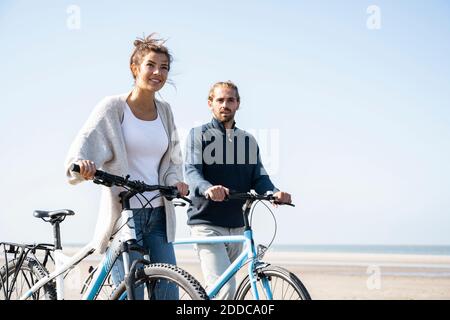Young couple walking with bicycles at beach against clear sky on sunny day Stock Photo