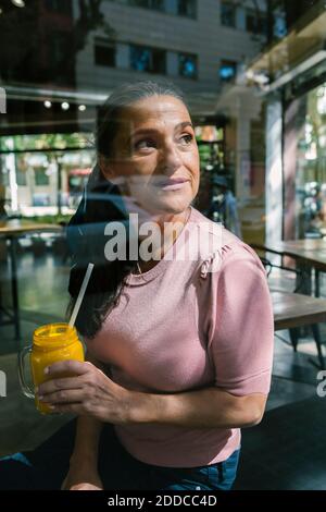 Thoughtful woman holding mason jar with juice while looking through window at cafe Stock Photo