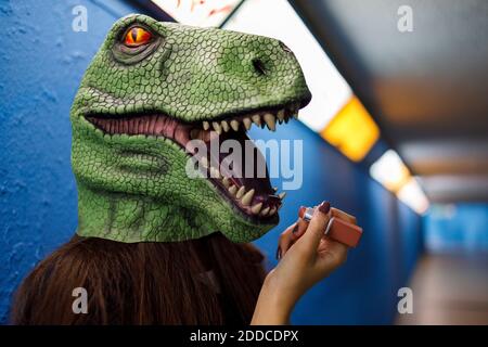 Woman applying lipstick while wearing dinosaur mask against blue wall Stock Photo