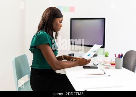 Businesswoman analyzing graph paper while sitting by desk at office Stock Photo