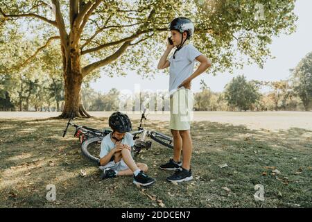 Boy talking on smart phone while brother holding hurt sitting in public park Stock Photo