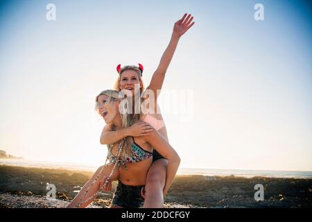 Happy young woman piggybacking female friend while enjoying weekend at beach during sunset Stock Photo