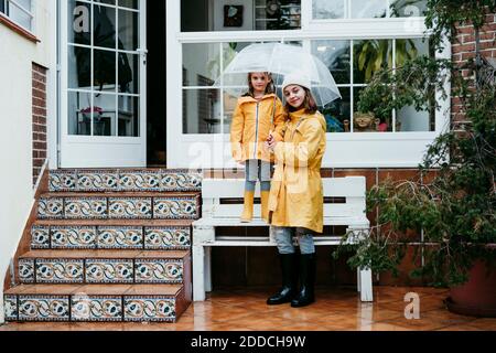 Smiling sister holding umbrella by sister standing on bench against home Stock Photo