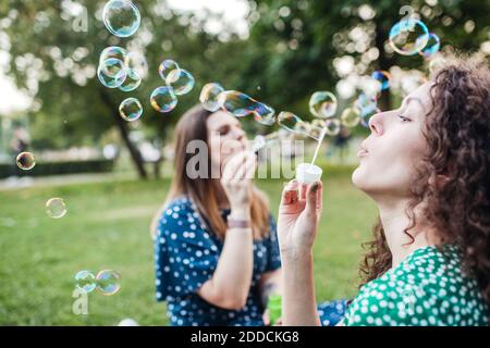 Beautiful female friends enjoying while blowing bubbles from wands at park Stock Photo