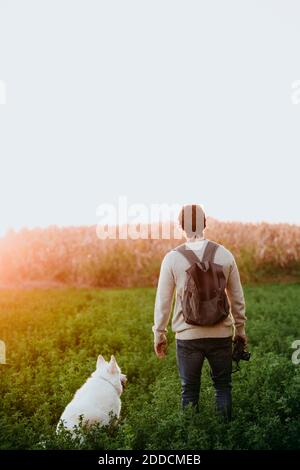 Man with backpack holding camera while sanding by dog at field Stock Photo