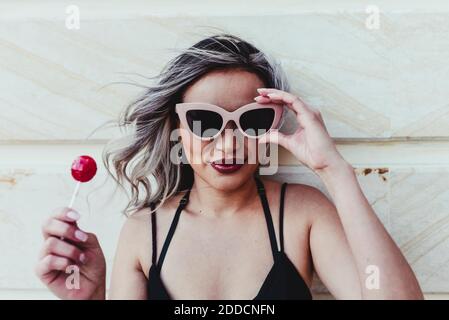 Beautiful woman wearing sunglasses while holding red lollipop against wall Stock Photo