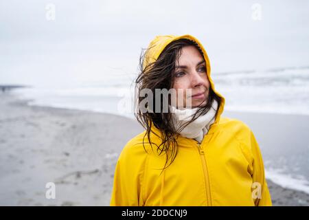 Thoughtful mature woman in yellow raincoat standing at beach against sky Stock Photo