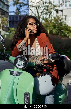 Young woman talking through smart phone while sitting on electric scooter and looking away Stock Photo