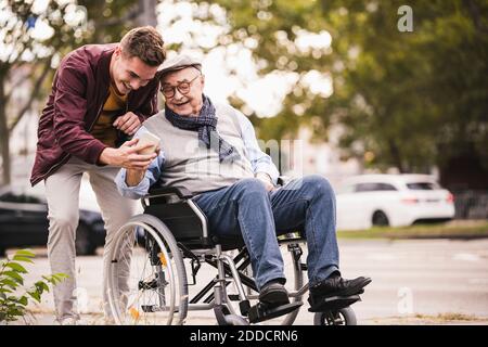 Senior man in wheelchair and his adult grandson looking together at smartphone Stock Photo