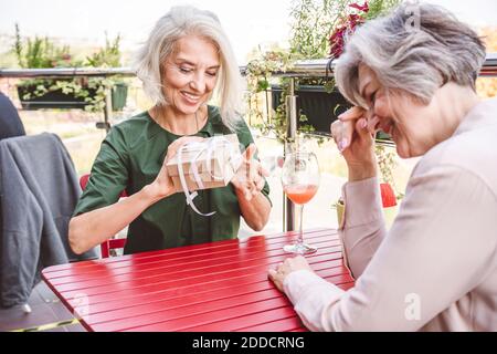 Smiling woman opening gift while sitting with female friend at restaurant Stock Photo