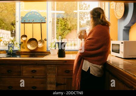 Senior woman in covered blanket looking through window while drinking coffee at home Stock Photo