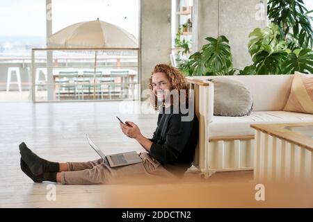 Smiling man using smart phone sitting with laptop on floor in living room Stock Photo