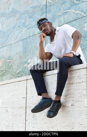 Young man with hand on chin sitting on retaining wall Stock Photo