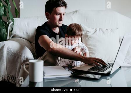 Father using laptop while son sitting on his lap at home Stock Photo