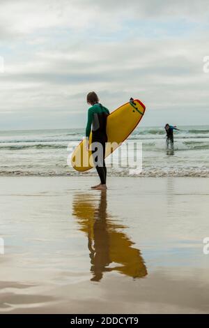 Teenage boy standing with surfboard while male friend standing in sea at beach Stock Photo