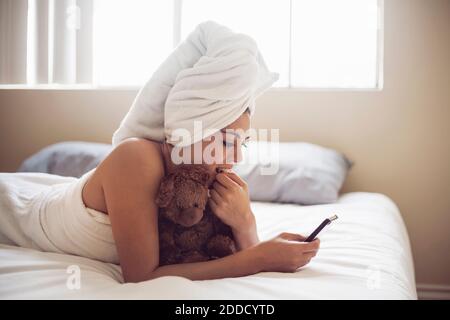 Young woman wrapped in towel using smart phone while lying on bed at home Stock Photo