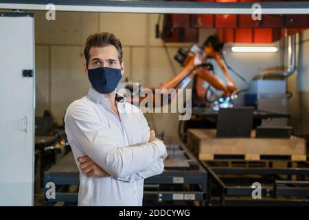Businessman wearing face mask standing with arms crossed against robotic arm equipment at factory Stock Photo