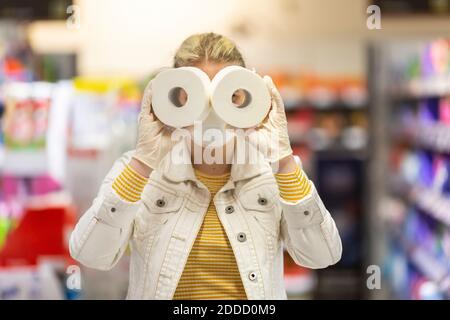 Teenage girl wearing protectice mask and gloves holding looking through holes of toilet rolls at supermarket Stock Photo