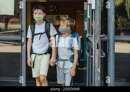 Brothers wearing protective face mask holding hands coming out of school building Stock Photo