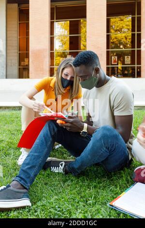 Male and female friends sitting on grass while studying together in university campus Stock Photo