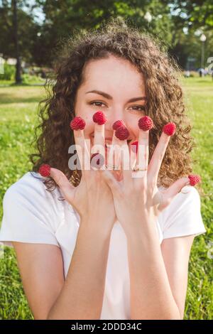 Playful young woman with raspberries on fingers at park during sunny day Stock Photo
