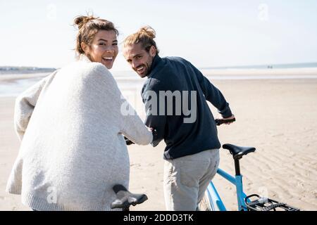 Happy young couple walking with bicycles at beach against clear sky on sunny day Stock Photo