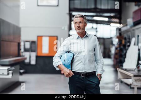 Thoughtful male engineer holding hardhat while standing in factory Stock Photo