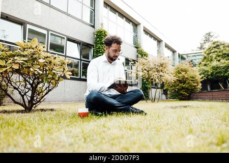 Businessman using digital tablet while sitting with coffee cup on grass against industry Stock Photo