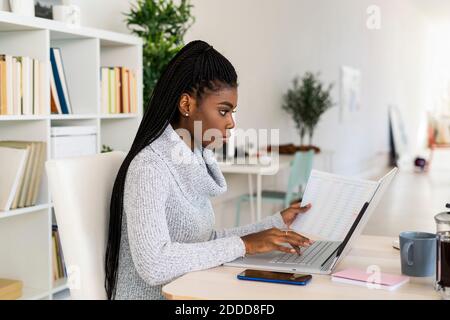 Female student concentrating while reading paper studying on laptop at home Stock Photo
