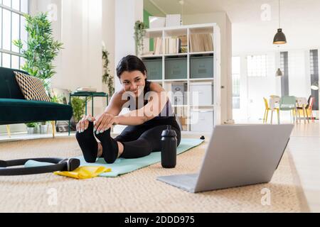 Smiling young woman looking away while doing stretching exercise sitting at home Stock Photo