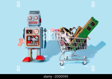 Studio shot of vintage robot toy standing by miniature shopping cart filled with electronic equipment Stock Photo