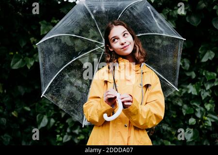 Thoughtful girl smiling while holding umbrella against leaf wall Stock Photo