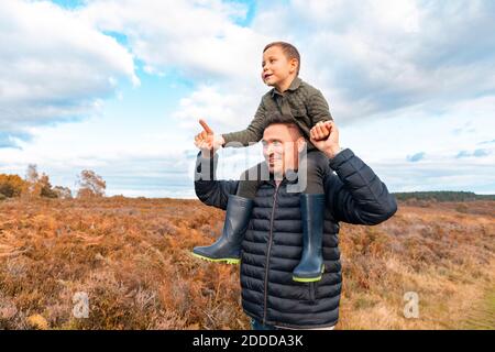 Father carrying son on shoulder standing in park against cloudy sky during autumn Stock Photo